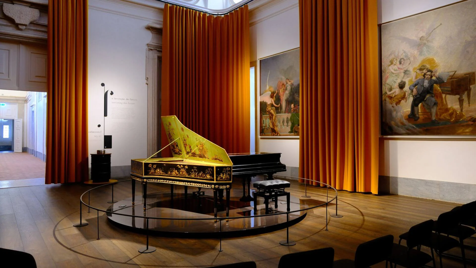Sala do Museu Nacional da Música com um cravo pintado e um piano de cauda num estrado circular, entre cortinas cor de laranja e grandes pinturas.
